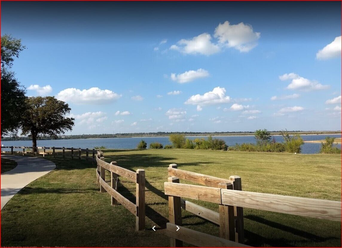 A wooden fence, a pathway, and a grassy area with a lake and trees in the background.
