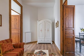A room with a wooden floor, a red sofa with a patterned cushion, a white cabinet with decorative elements, and a dining area with a table and chairs.