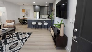 A modern kitchen with dark cabinets, a central island with stools, a flat-screen TV mounted above the island, and a dining area with a patterned rug and chairs. - Trendy & Oh-So Contemporary (Scottsdale)