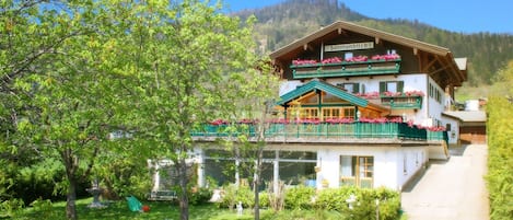 A traditional Alpine-style building with a balcony adorned with flowers, surrounded by lush greenery and a mountain backdrop.