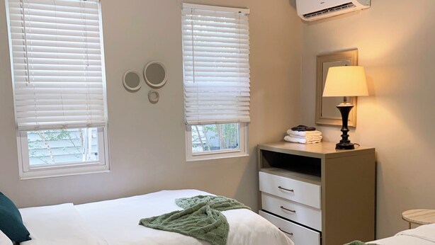 A hotel room with two beds, a bedside table, a lamp, a dresser, and an air conditioning unit.