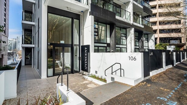 A modern building with a glass facade, numbered 106, featuring a gated entrance and a small outdoor area with plants. - Relaxing City Center Studio Close to Queen St (Auckland)