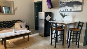 Eine moderne Küche mit Kühlschrank, einem Esstisch mit zwei Stühlen, einer Couch mit Blumenkissen und einem wandmontierten Fernseher.