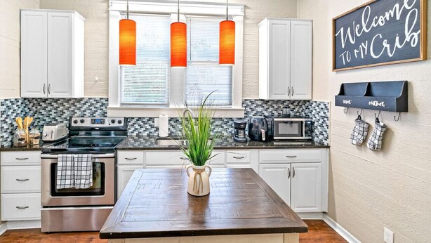 A kitchen with white cabinets, a wooden island, a blackboard with 'Welcome to My Crib' written on it, and hanging kitchen towels.