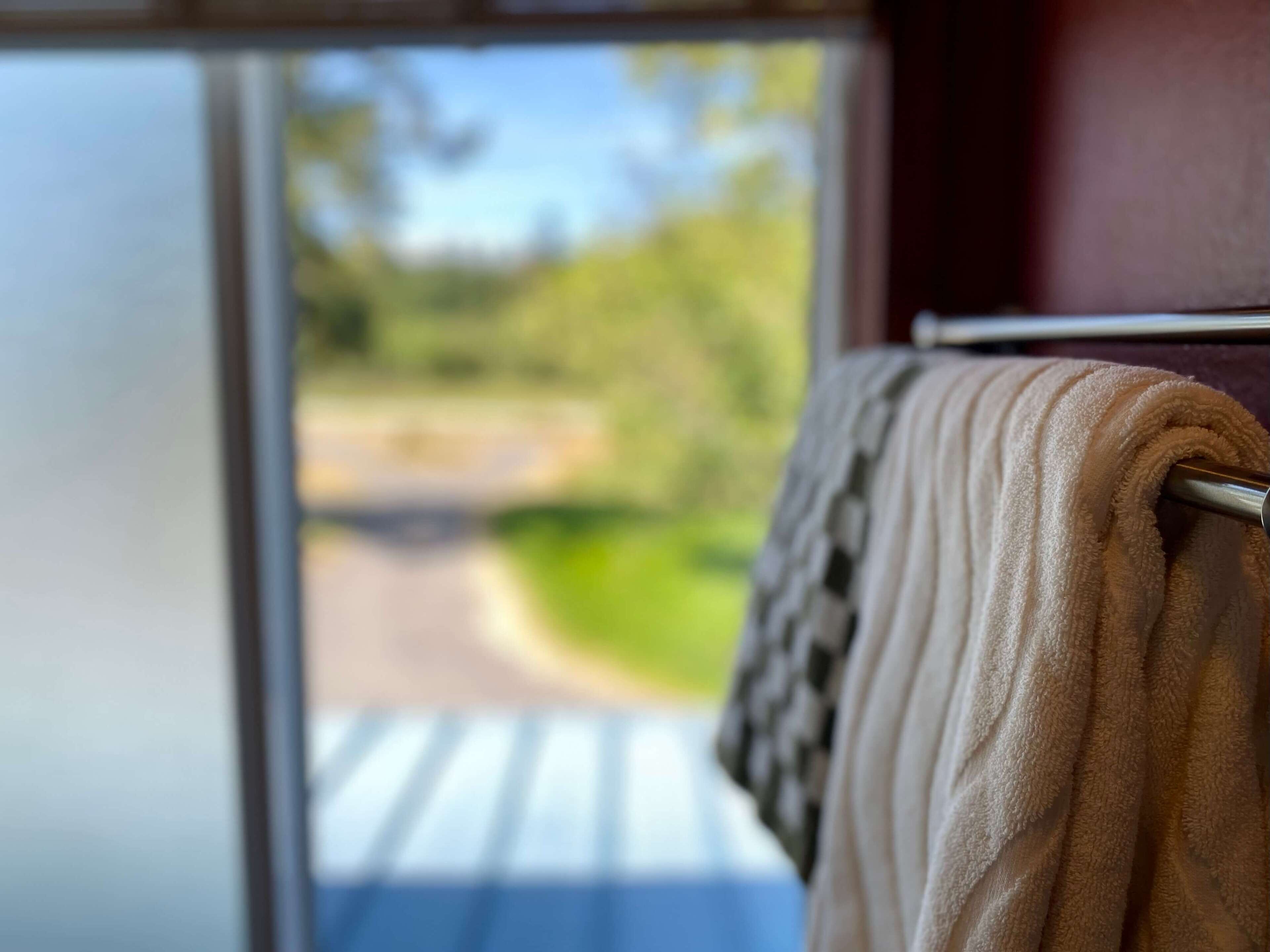 A window with a towel hanging on a metal bar, a view of a road, and greenery outside.