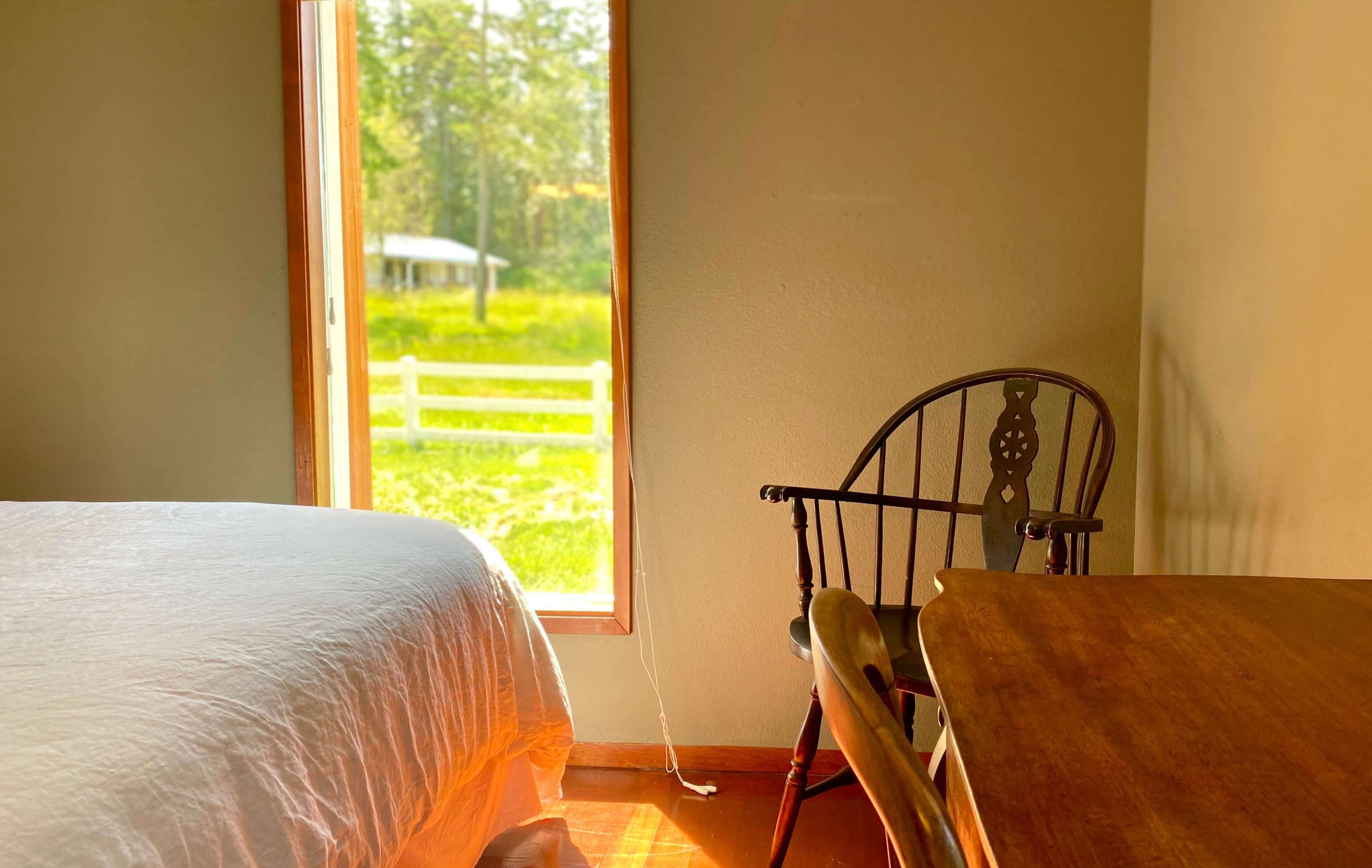 Quarto com cama, mesa de madeira e cadeira. Janela com vista para paisagem verde e cerca branca.