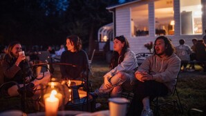 A group of people sitting around a campfire at night, with a house in the background. - Saturn's Return (Friday Harbor)
