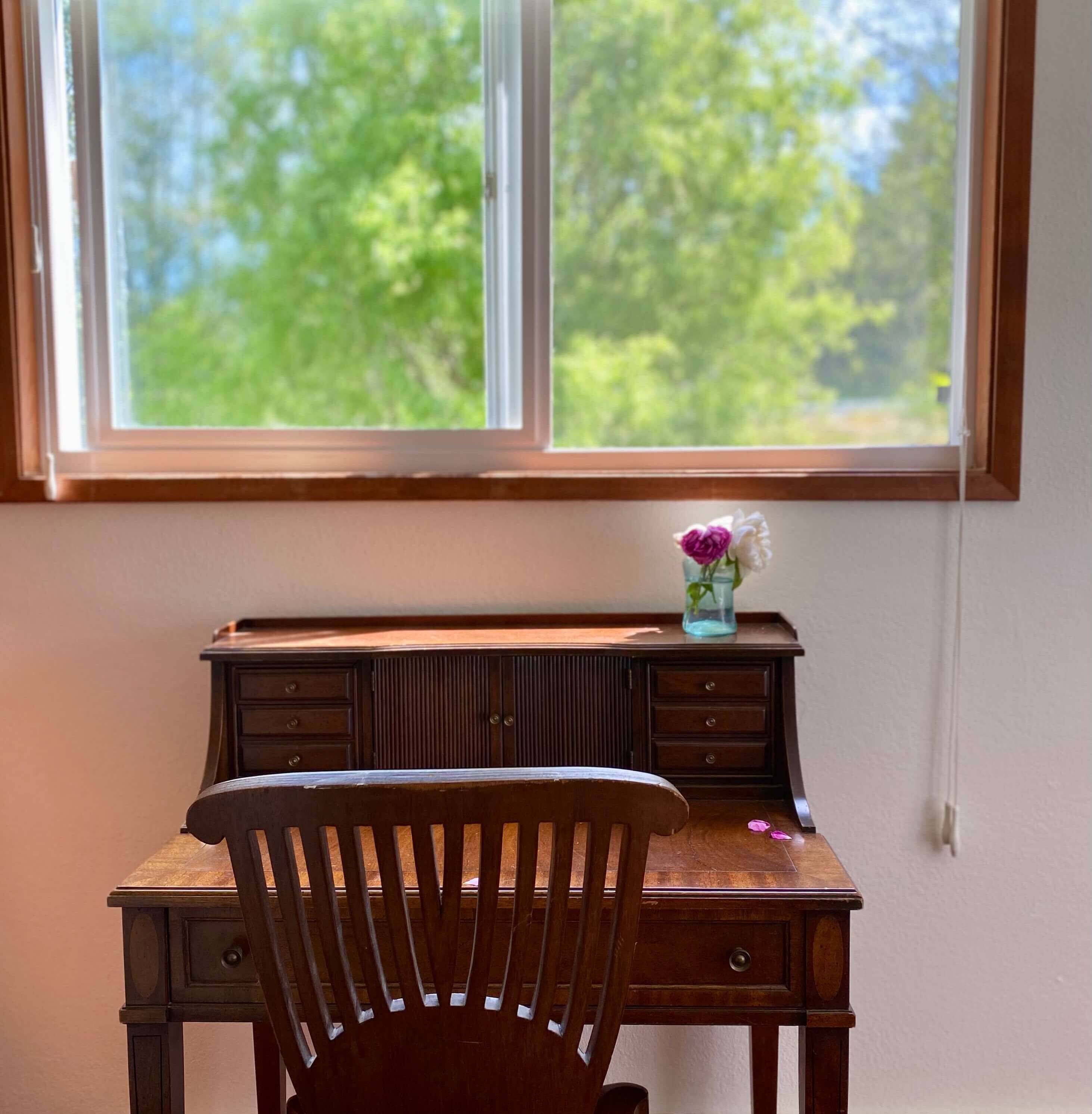 Un escritorio de madera con una silla frente a una ventana que da a un paisaje de árboles.