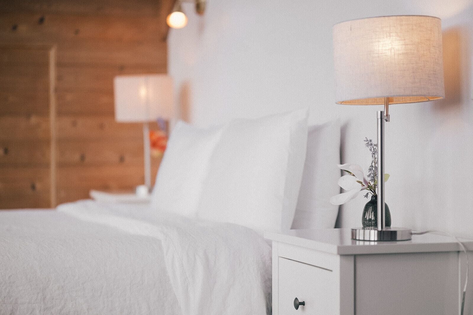A neatly made bed with white linens, a bedside table with a lamp and a vase of flowers, and a wooden headboard.