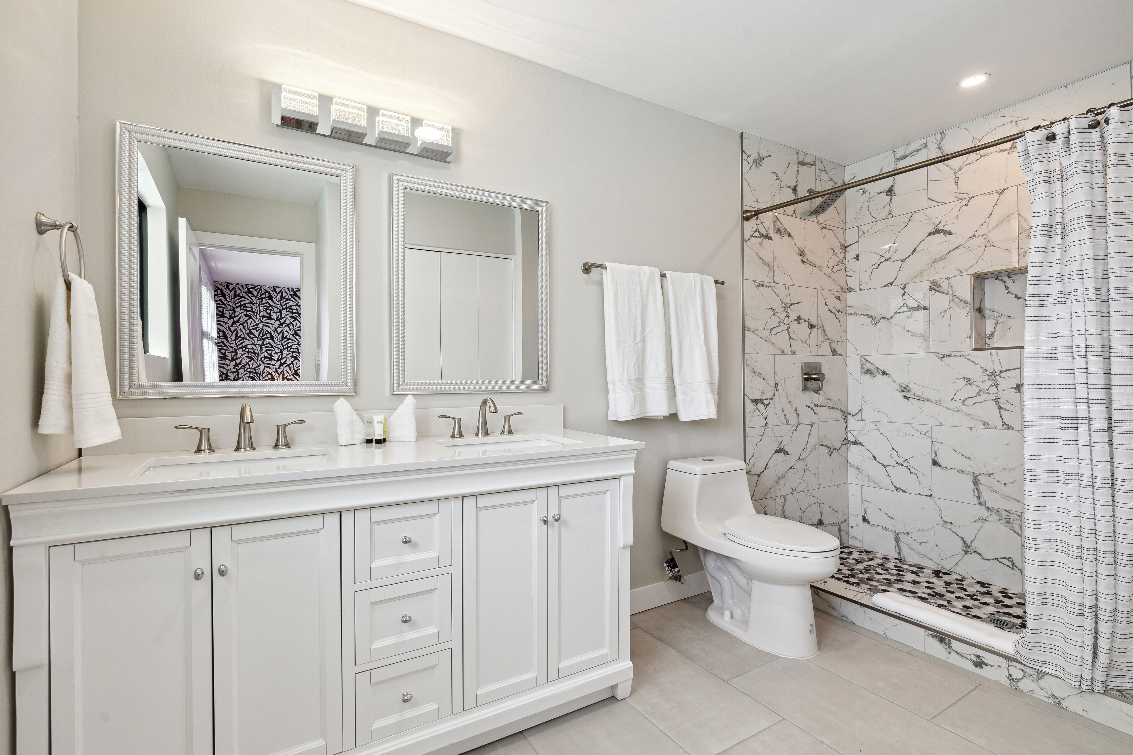 Un baño moderno con un espejo grande, dos lavamanos, un inodoro y una ducha a ras de suelo con pared de mármol.