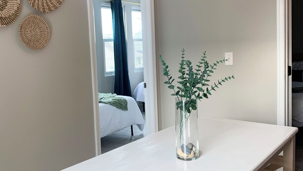 Comedor con una mesa blanca, dos sillas de madera, un florero con planta y decoración en la pared.