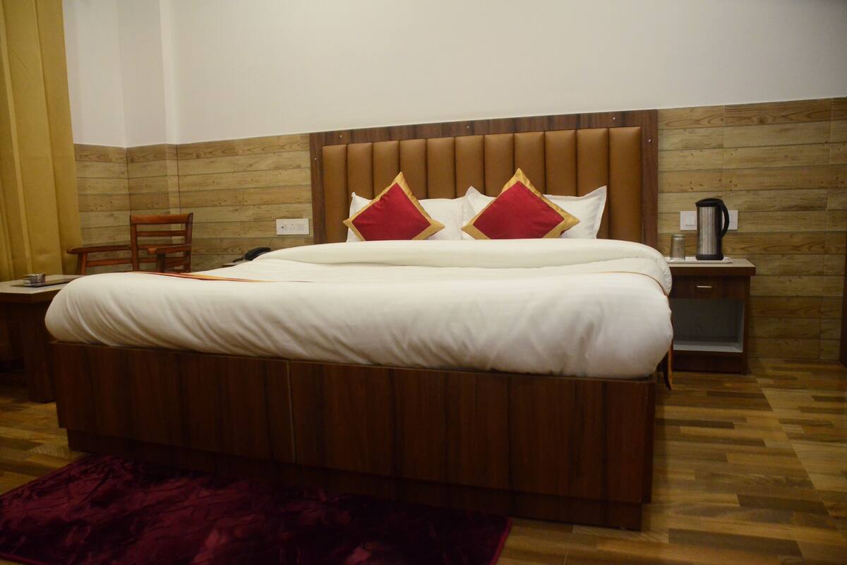 A hotel room with a wooden headboard, a bed with white bedding and red pillows, a wooden nightstand with a coffee maker, and a chair.
