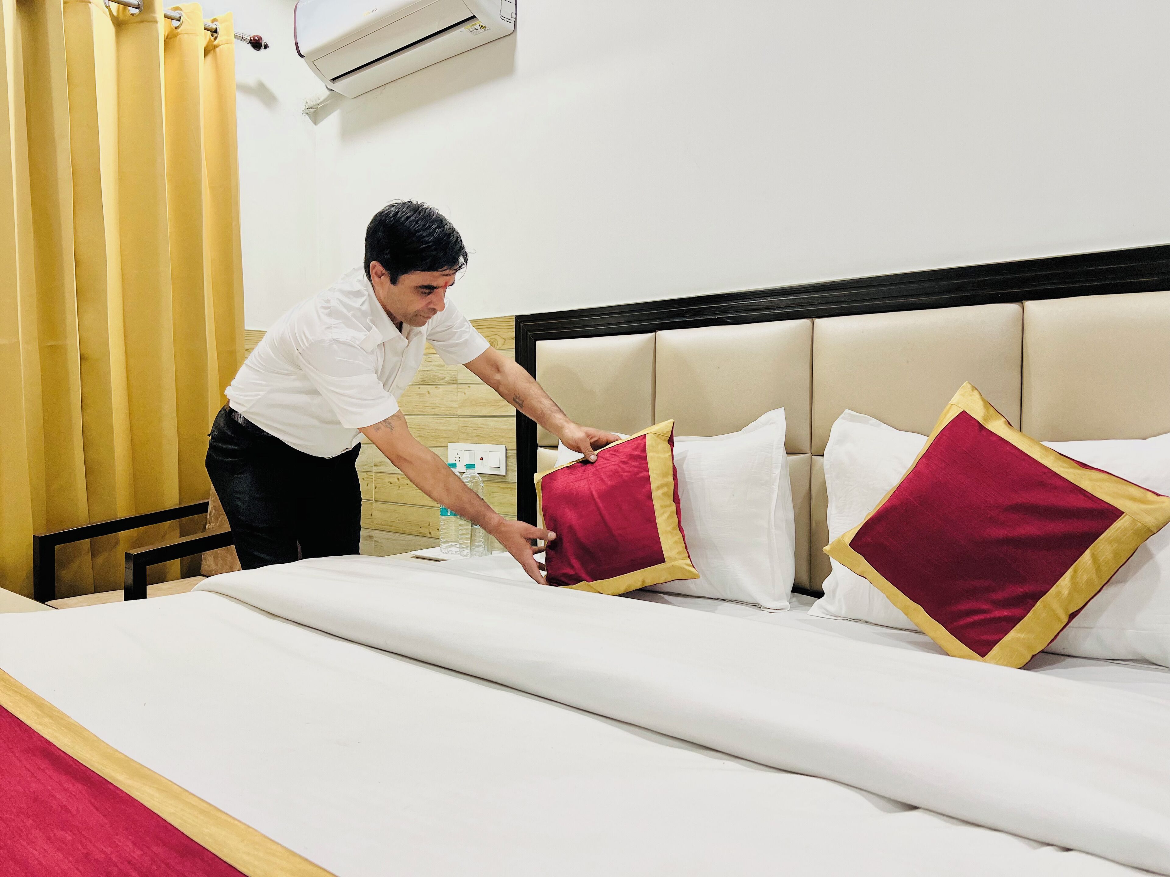 A hotel room with a bed, pillows, a bedside table, and an air conditioning unit.