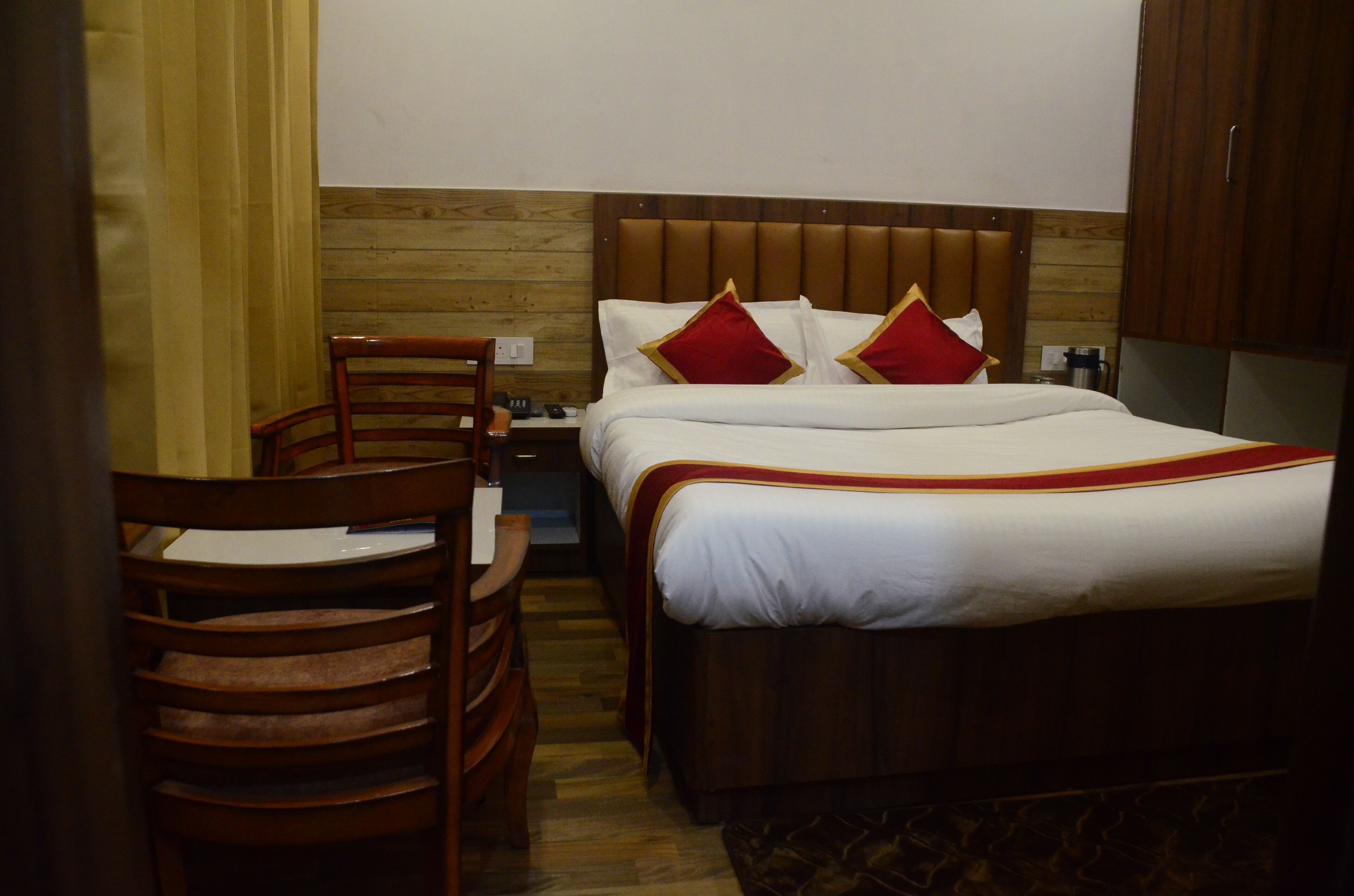 Habitación de hotel con cabecera de madera, una silla de madera, una mesita de noche y una cama con ropa de cama blanca y almohadas rojas.