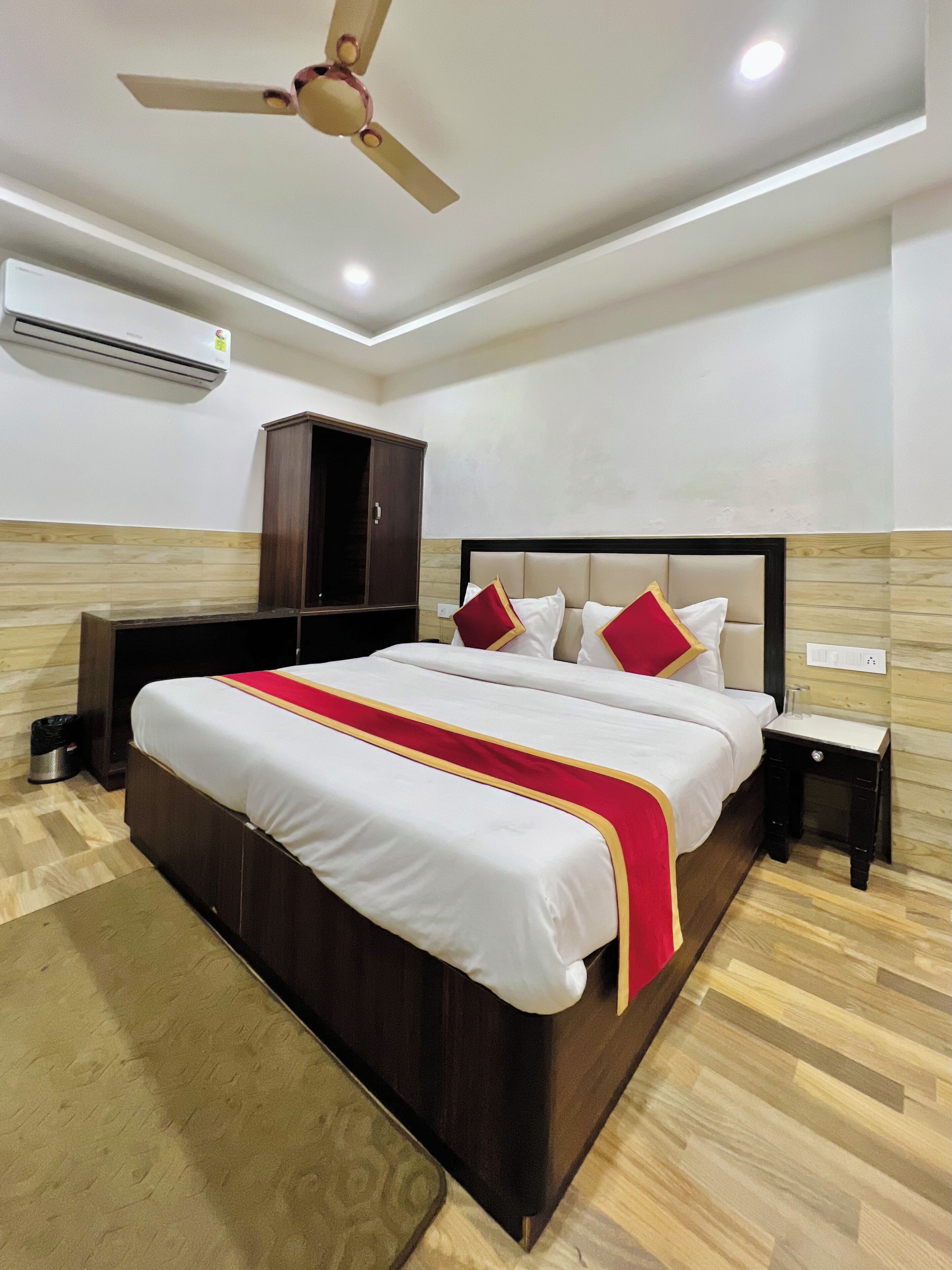A hotel room with a large bed, a ceiling fan, an air conditioning unit, a wooden wardrobe, and a bedside table.
