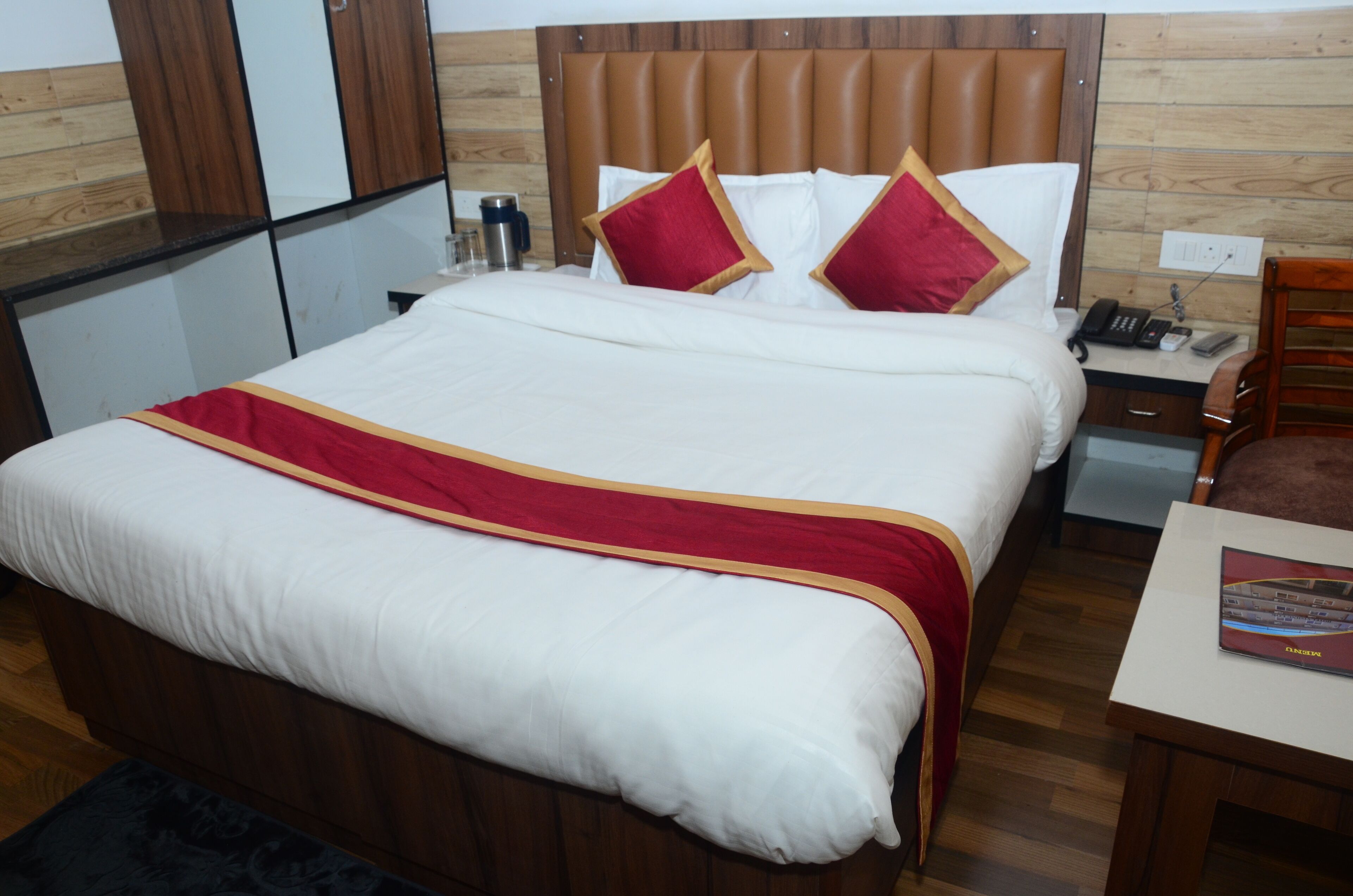 Habitación de hotel con una cama grande, cabecera de madera, mesita de noche con lámpara y una pequeña mesa con una revista.