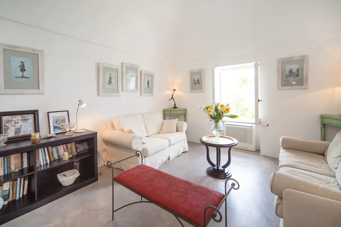 Ein Wohnzimmer mit einem weißen Sofa, einer roten Bank, einem Bücherregal und einem kleinen Tisch mit einem Blumenstrauß.