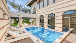 A swimming pool with lounge chairs and umbrellas, surrounded by a wooden deck and a building with arched windows. - Maison Privee - Majestic Resort Villa with Private Pool on The Palm (Dubai)