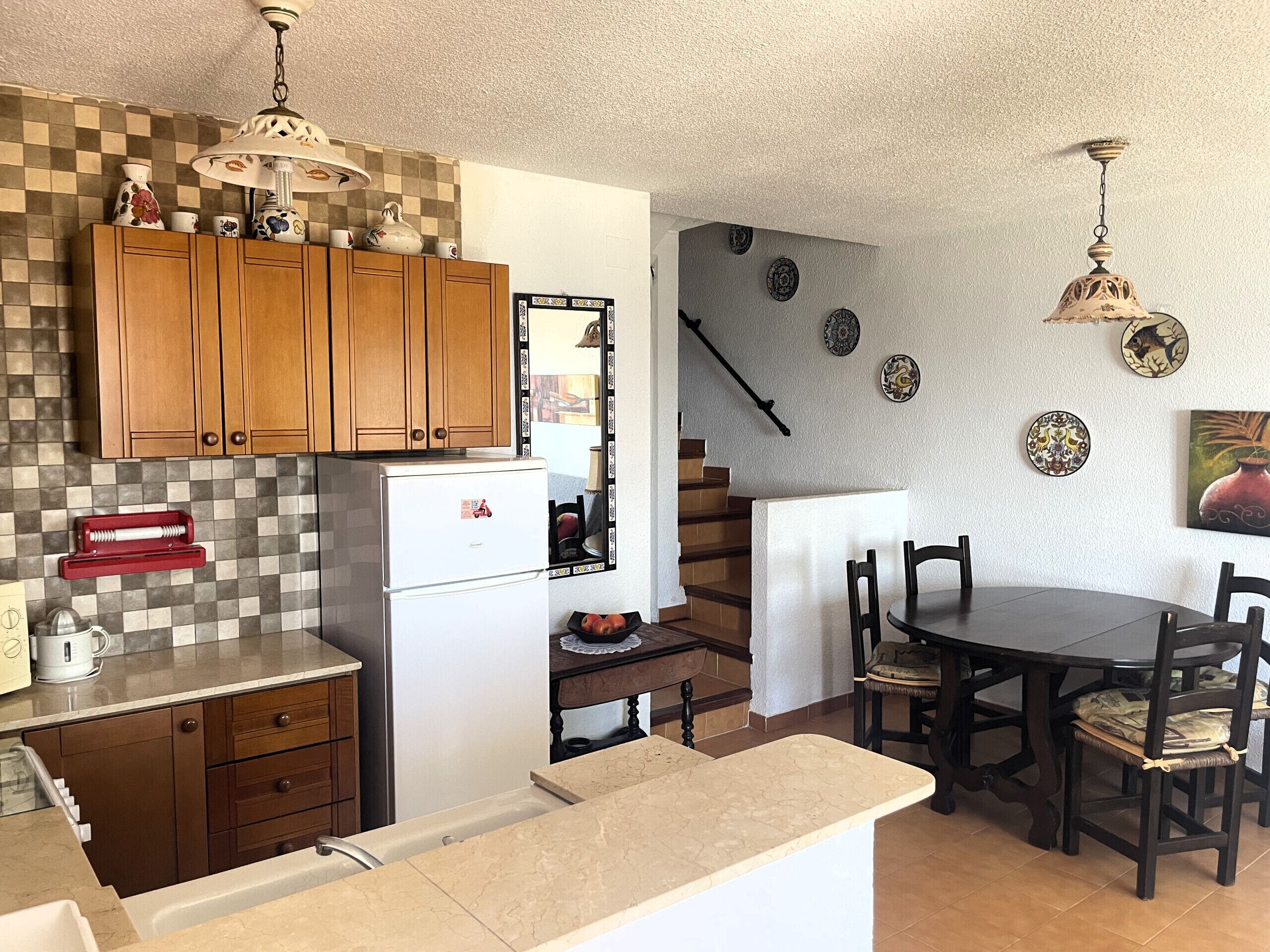 A kitchen with wooden cabinets, a refrigerator, a dining table with chairs, and a staircase leading to another level. Decorative plates and a wall-mounted light fixture are also visible.