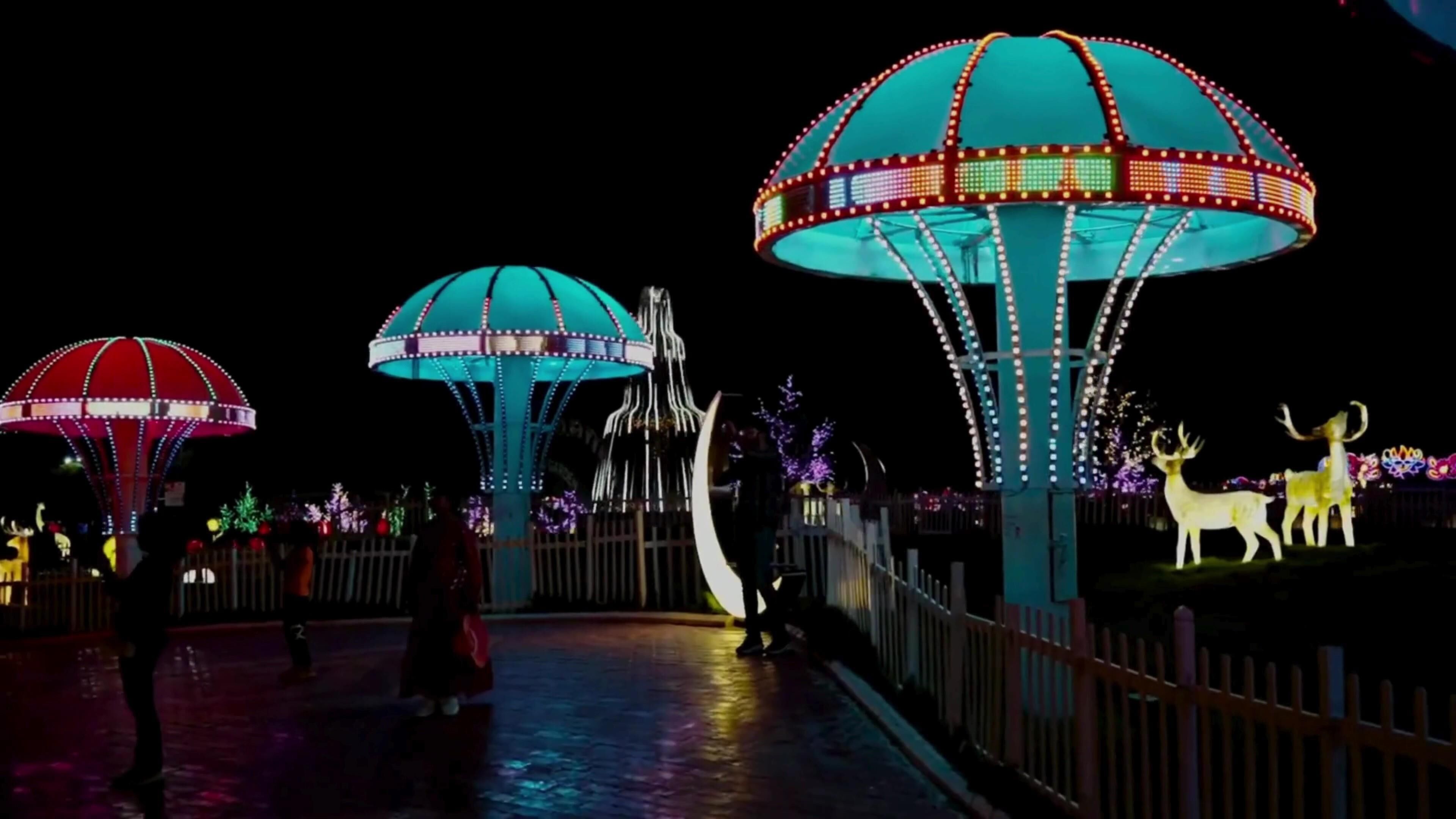 A nighttime scene with illuminated structures, including large domed kiosks and a crescent-shaped feature, surrounded by a fence and decorative elements like deer statues.