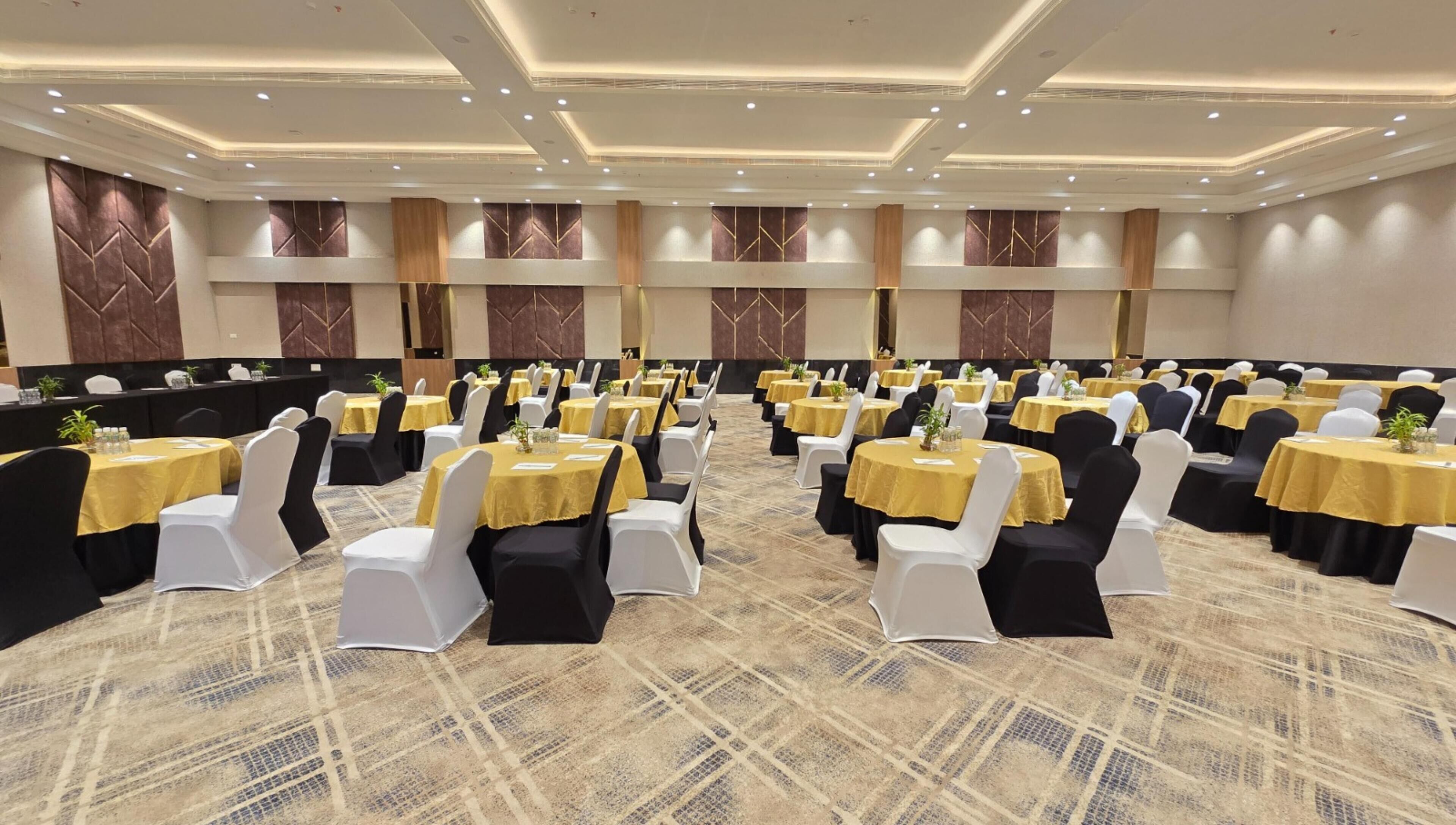 Un salón de banquetes con mesas redondas, sillas blancas y negras, y manteles amarillos.
