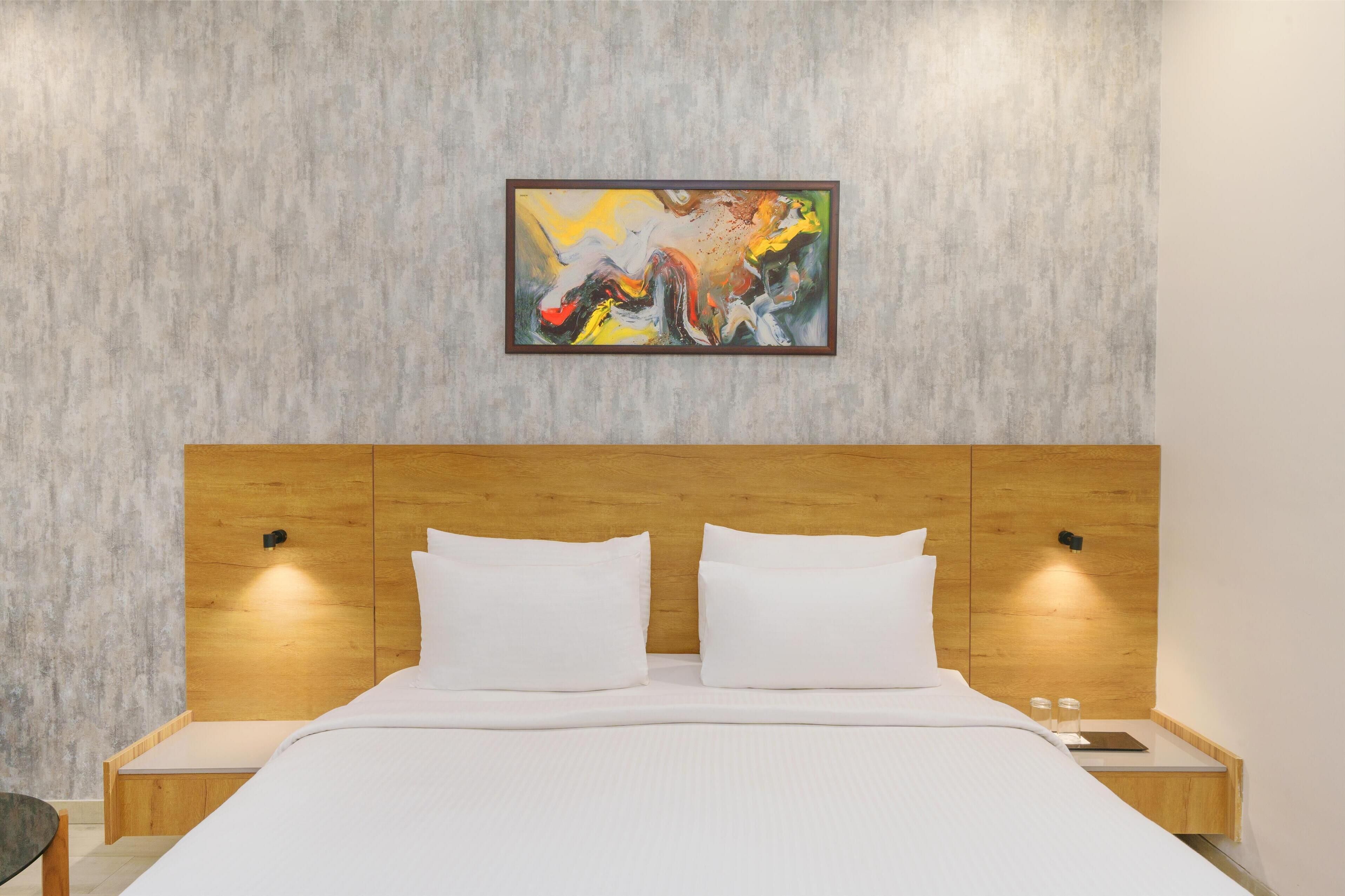 A modern hotel room with a wooden headboard, white bedding, and a colorful abstract painting on the wall.
