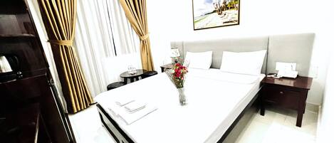 A hotel room with a large bed, a bedside table with a lamp, a nightstand with a phone, a small table with a vase of flowers, and a painting on the wall.