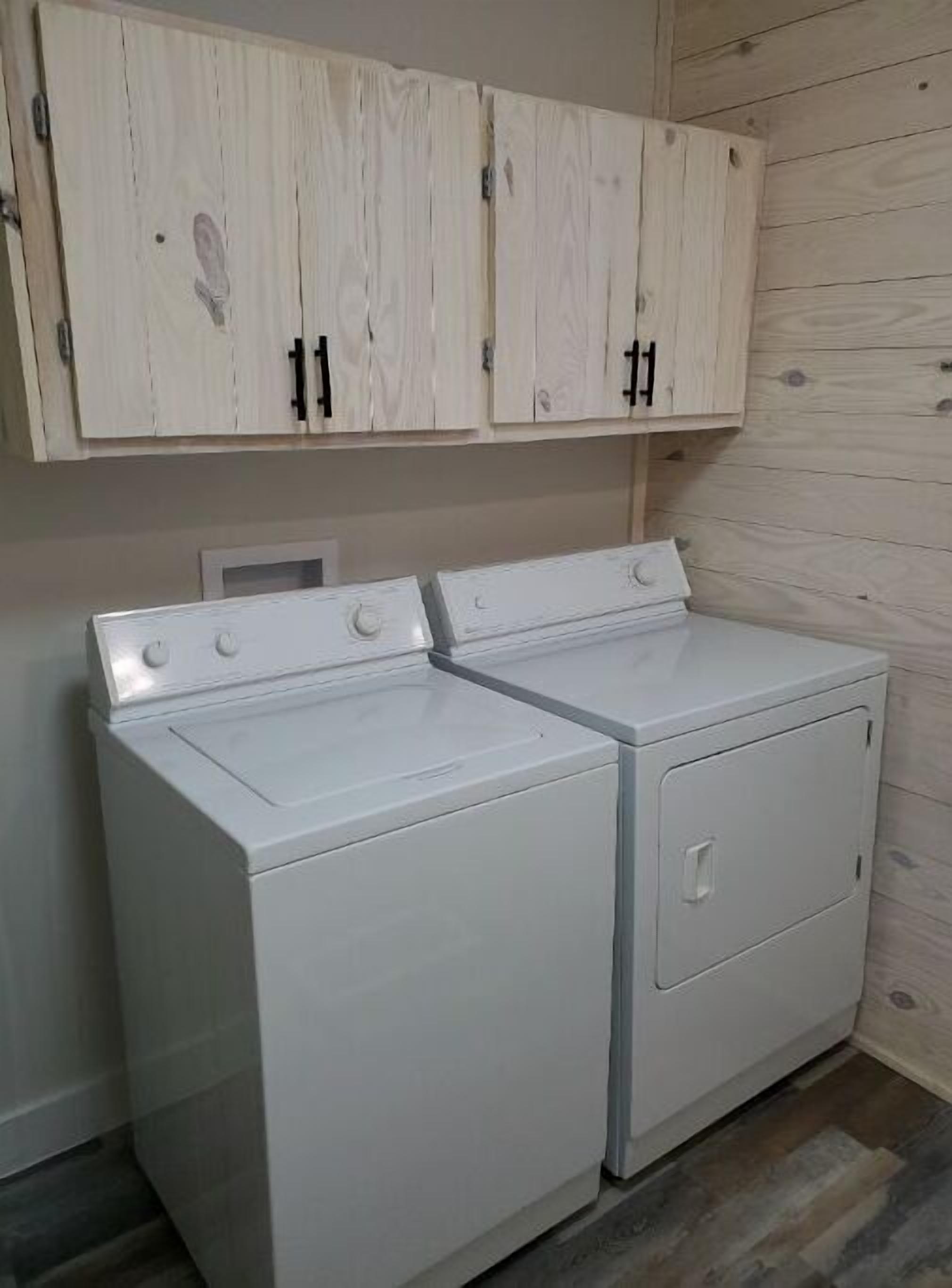 Une buanderie équipée de deux machines à laver et de deux sèche-linge à chargement frontal, tous deux de couleur blanche, avec des armoires en bois au-dessus et un plancher en bois.