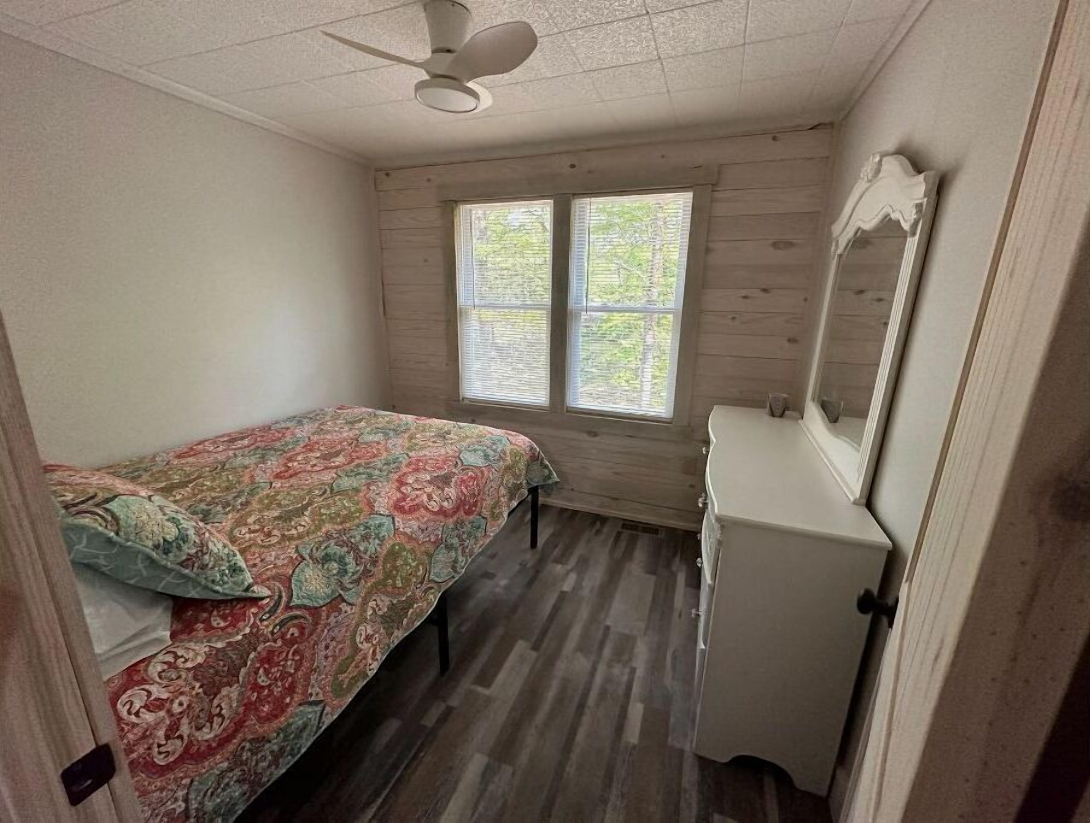 Une chambre à coucher comprenant un lit, une commode, un ventilateur de plafond et deux fenêtres.