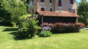 Um edifício de dois andares com telhado de telhas, rodeado por vegetação exuberante e plantas floridas.