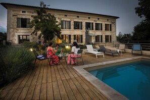 A wooden deck with a pool, outdoor seating, and a stone building in the background.