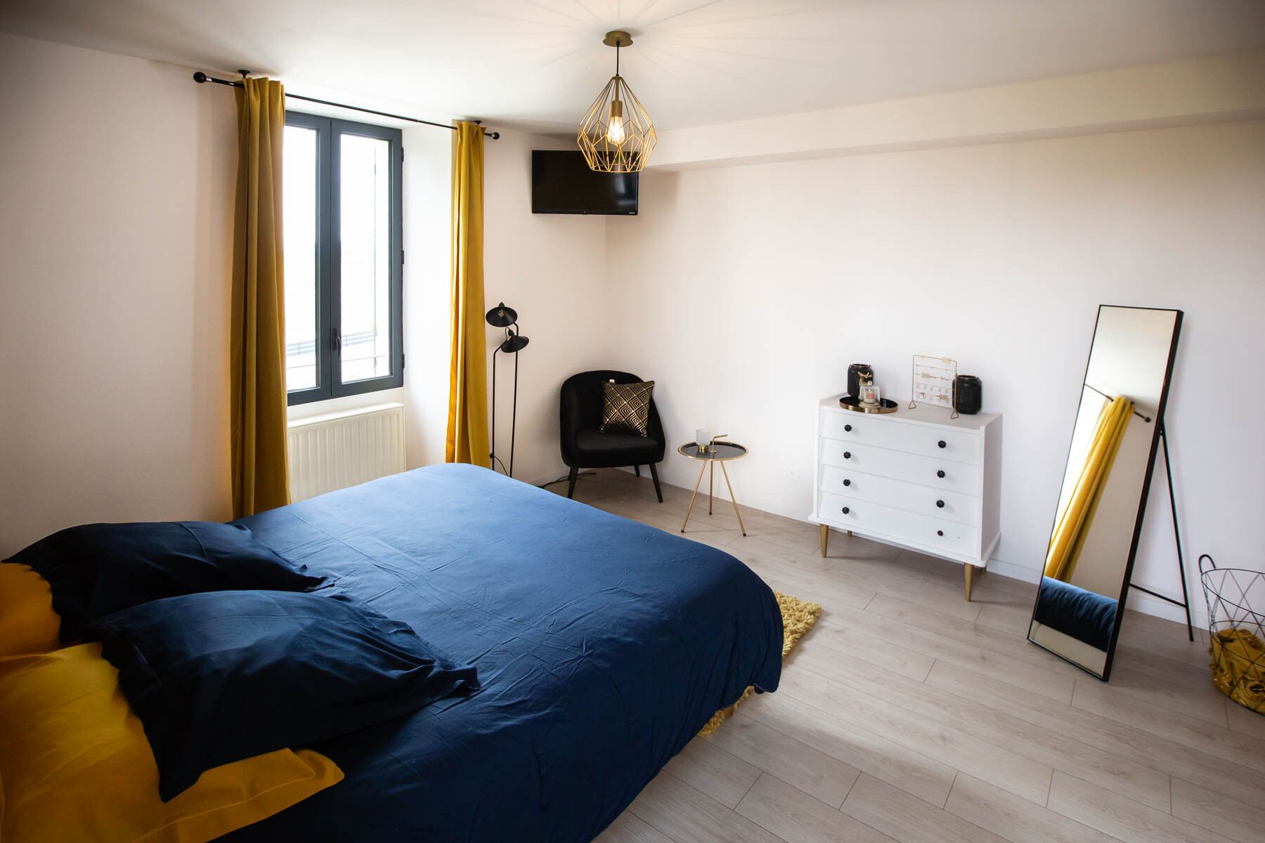 A bedroom with a bed, a dresser, a chair, a floor lamp, a mirror, and a TV.