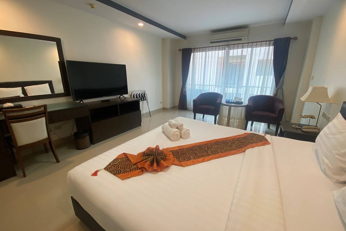 A hotel room with a bed, a desk with a TV, two chairs, a small table, and a window with curtains.