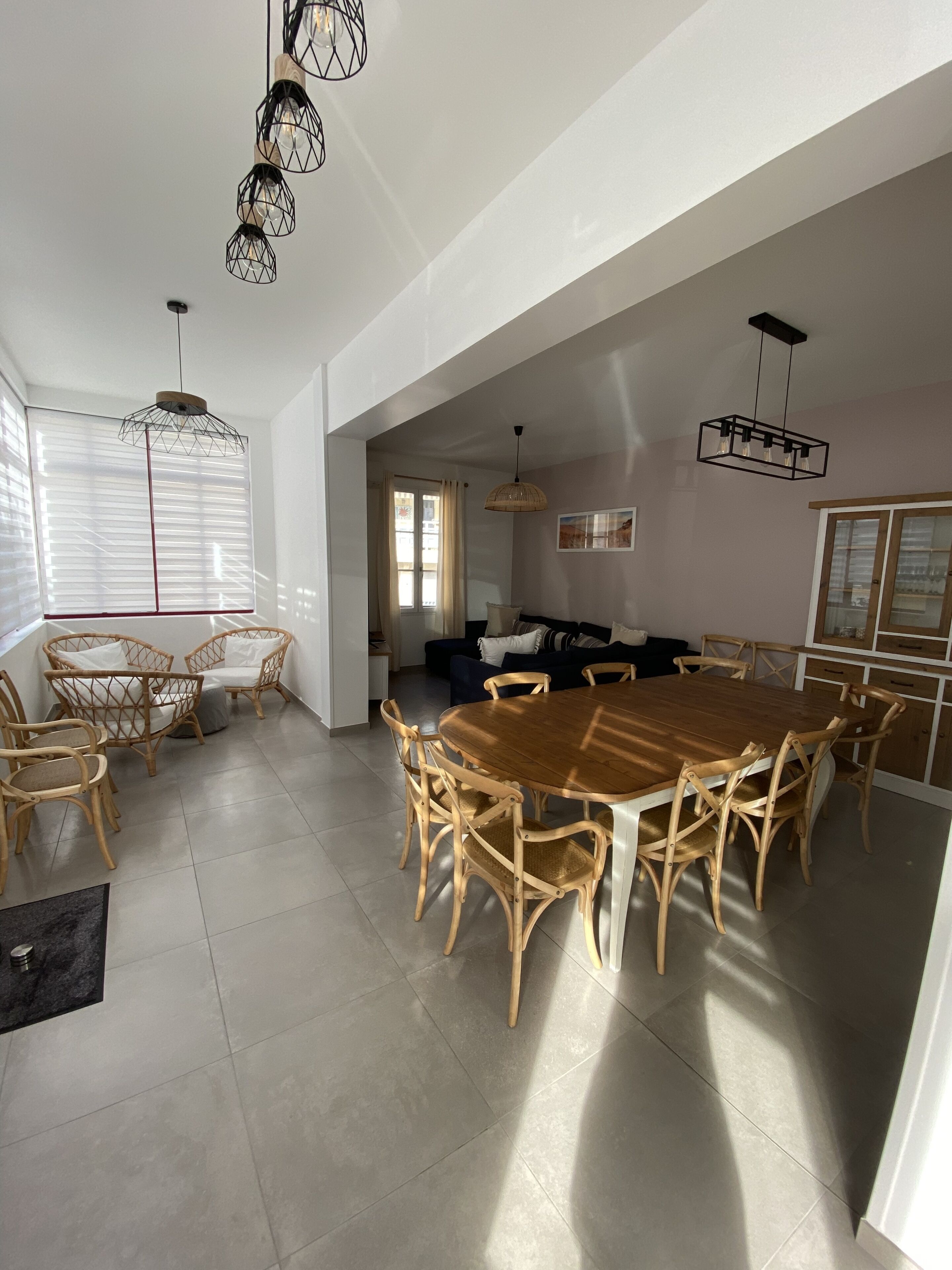 Un vaste espace de salle à manger avec une grande table en bois entourée de chaises en osier, un luminaire suspendu moderne et une vue sur la cuisine et le salon.