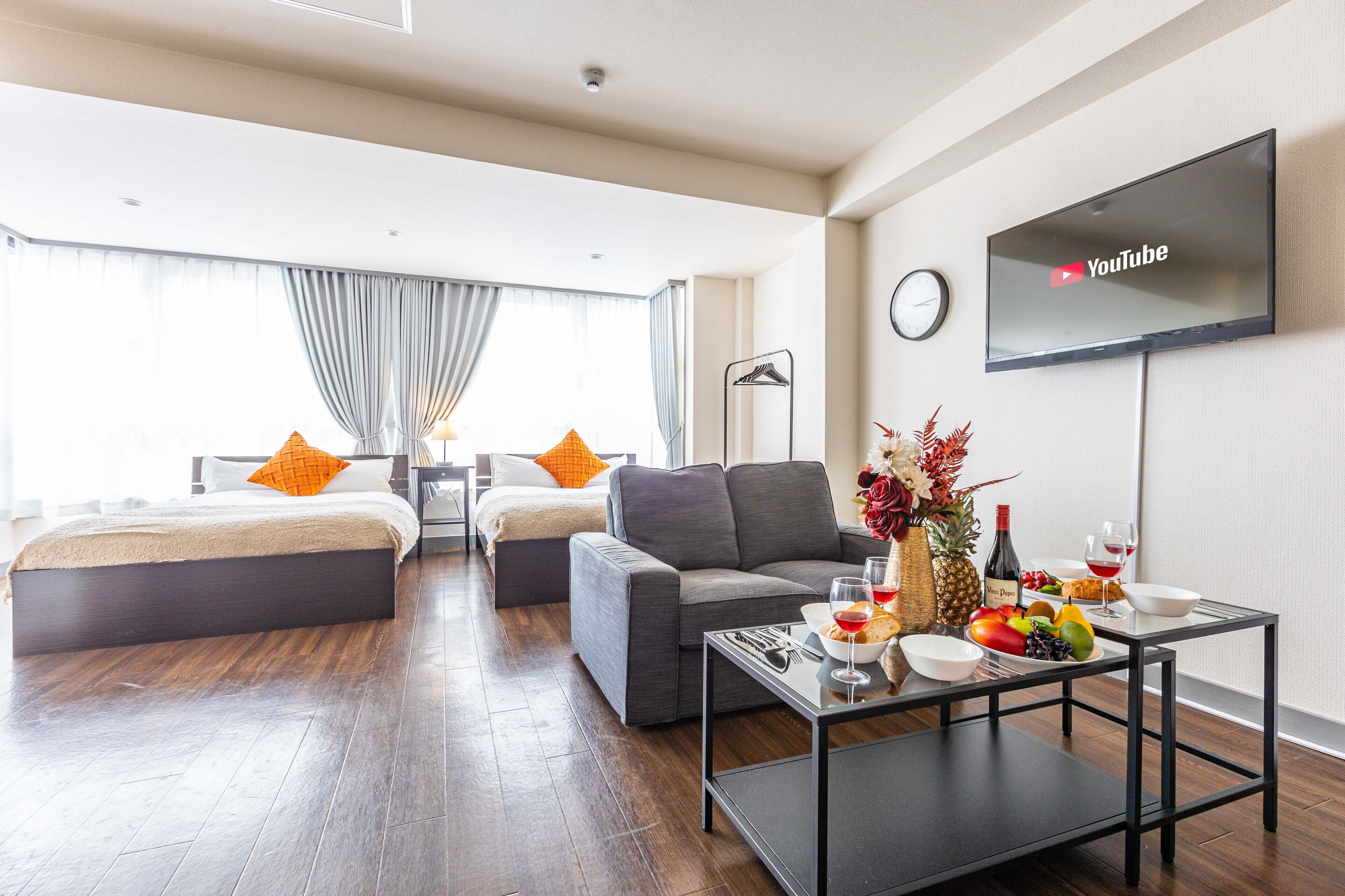 Une chambre d’hôtel moderne équipée d’un canapé, de deux lits, d’un téléviseur à écran plat, d’une table à manger avec des fruits et du vin, et d’une horloge murale.