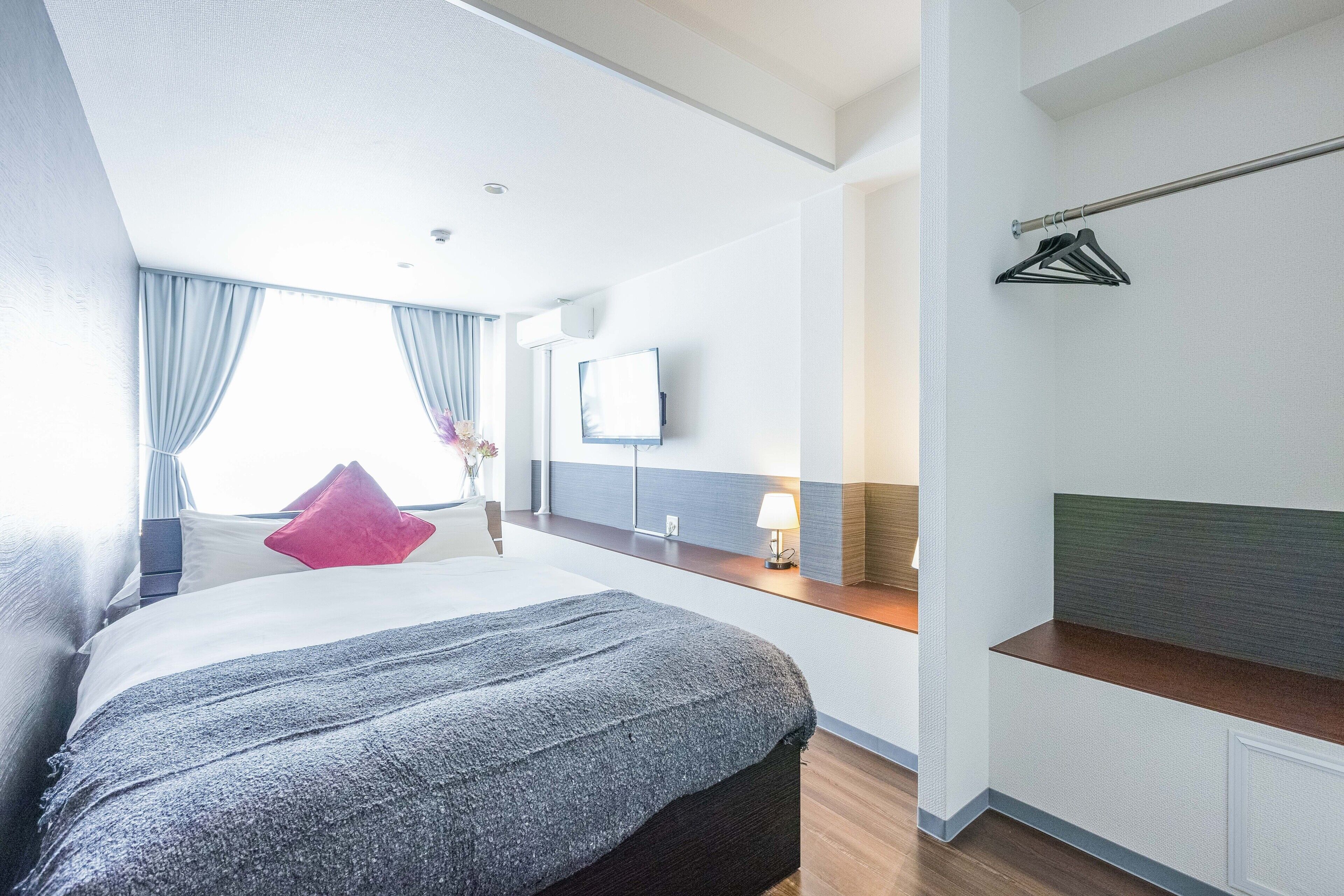 A modern hotel room with a large bed, a bedside table, a TV, and a window with curtains.