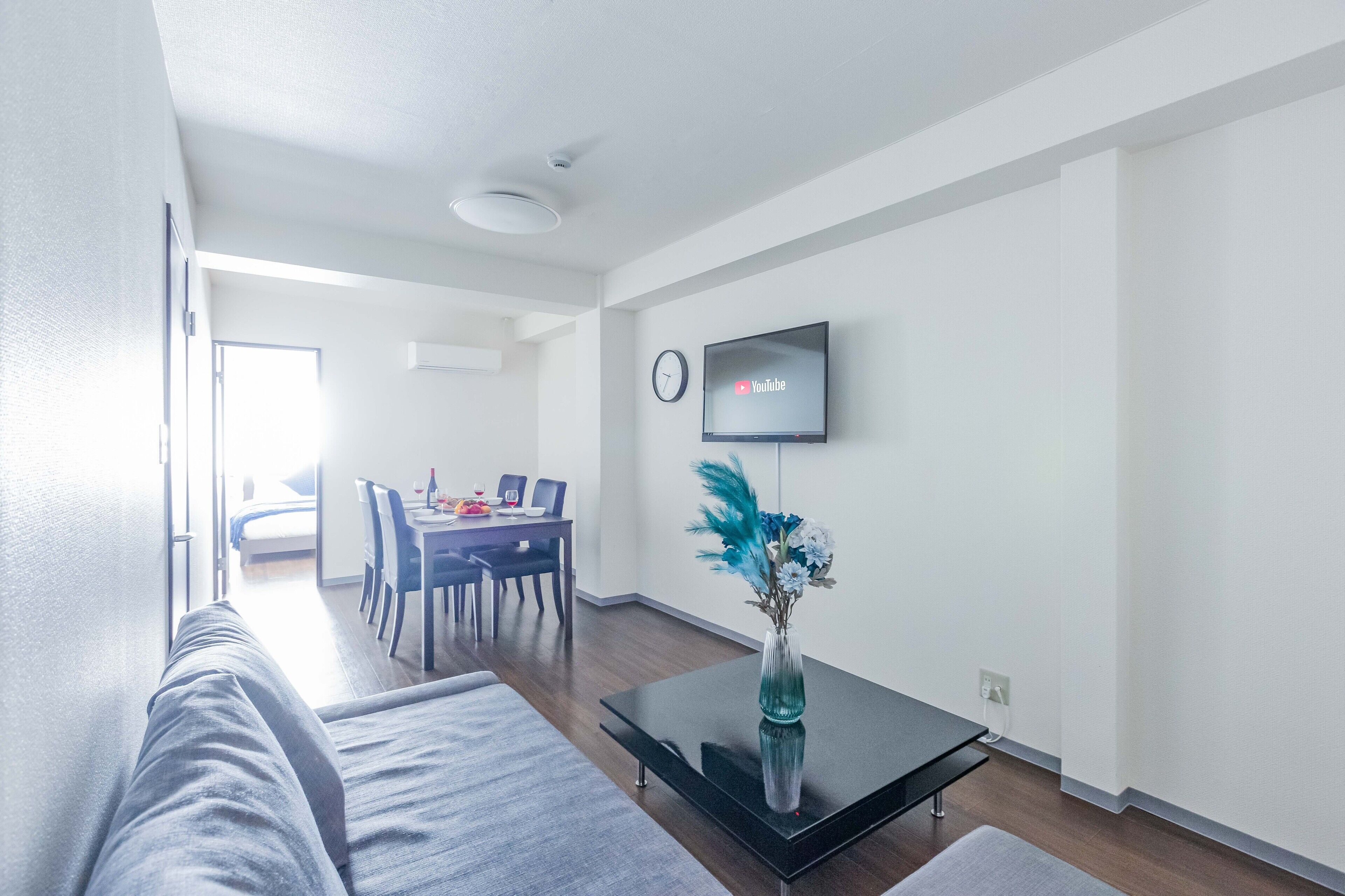Een moderne woonkamer met een bank, een salontafel met een vaas, een flatscreen-tv aan de muur en een eethoek op de achtergrond.