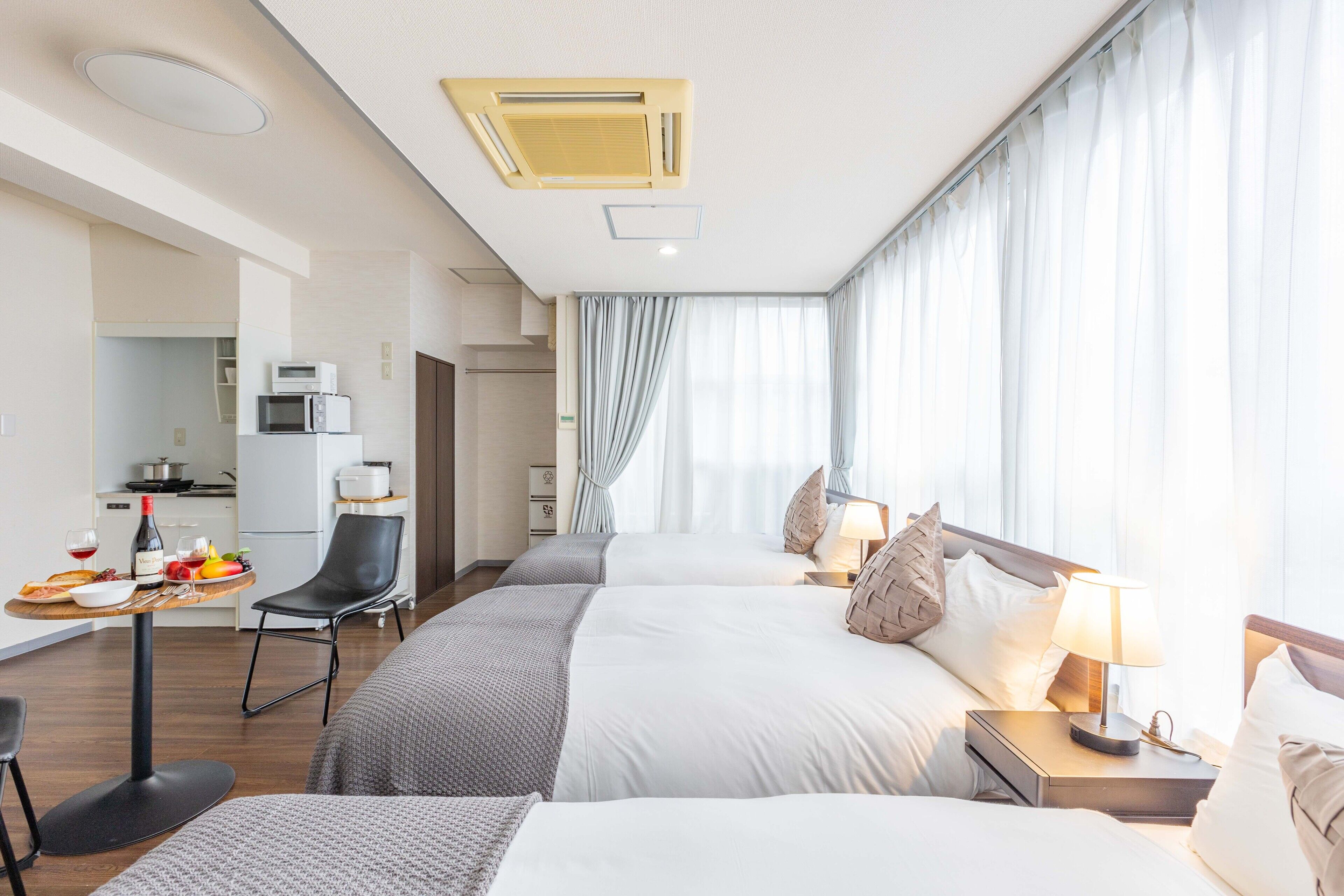 Une chambre d’hôtel avec deux lits, un coin repas comprenant une table et des chaises, une kitchenette et de grandes fenêtres avec des rideaux.