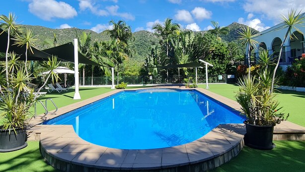 Una piscina rectangular con un borde perimetral, rodeada de césped artificial verde, palmeras y un paisaje montañoso de fondo.
