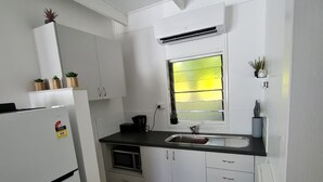 Cocina compacta con armarios blancos, encimera negra, microondas integrado, refrigerador, ventana con persianas y una pequeña planta en maceta sobre la encimera.