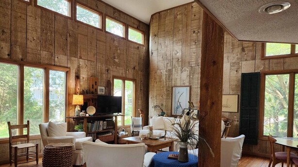 Un ampio soggiorno con pareti in legno, ampie finestre e un'accogliente area salotto con televisione, libri e piante decorative.