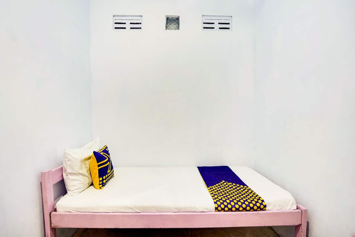 Una cama individual con colchón blanco, estructura rosa y dos almohadas con estampado azul y amarillo. Hay dos pequeñas rejillas en la pared sobre la cama.