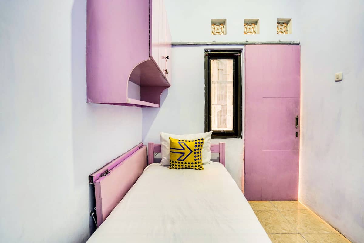 Una habitación pequeña con cama individual, en tonos rosa y blanco, una ventana y una puerta.
