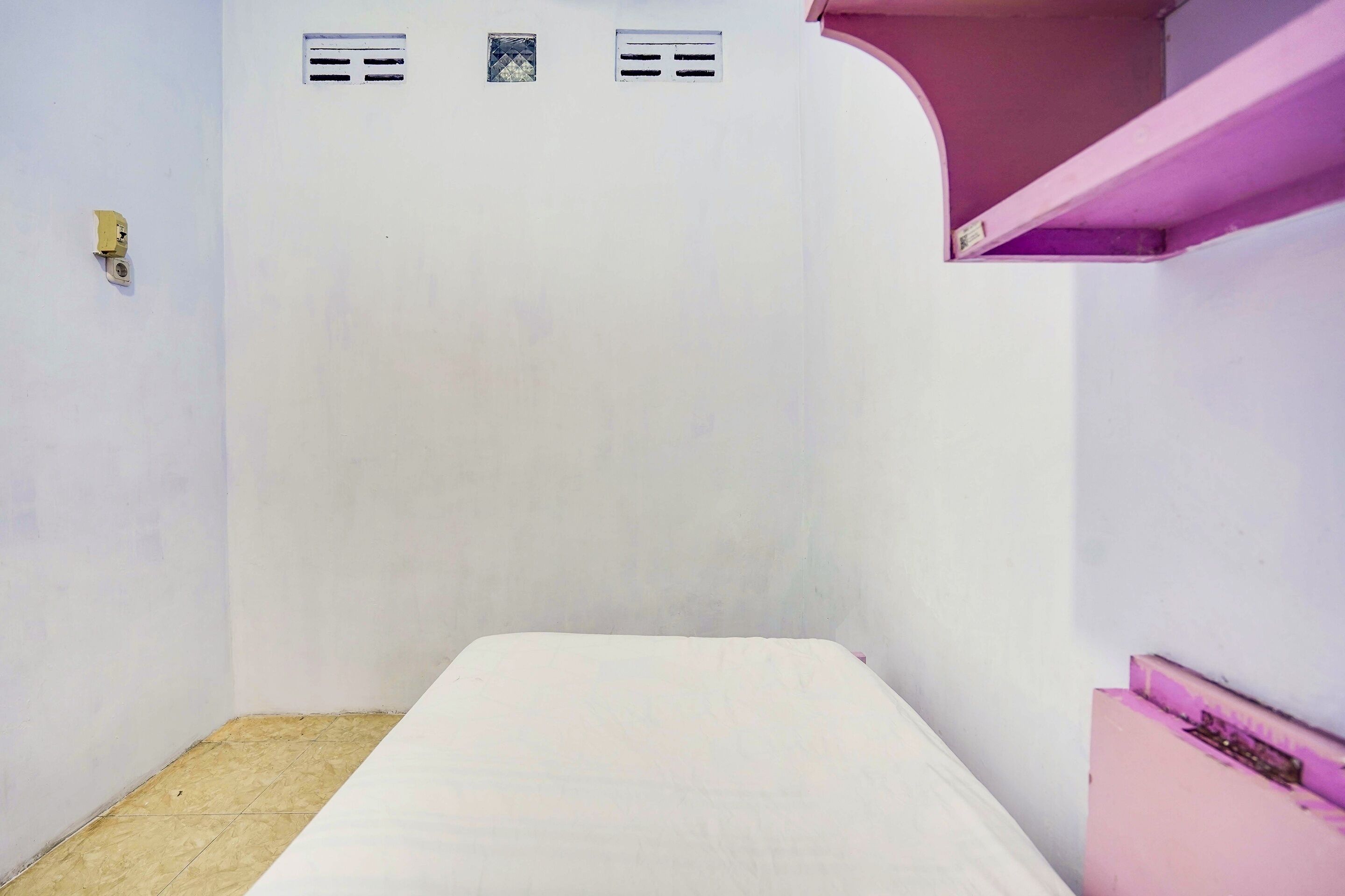A small, sparsely furnished room with a single bed, a pink shelf, and two air vents on the wall.