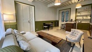 Ein Wohnzimmer mit einer Couch, einem Couchtisch und einem Sessel. Es gibt einen Essbereich mit Tisch und Stühlen. Im Hintergrund ist eine Küche mit einer Arbeitsplatte und Regalen zu sehen.