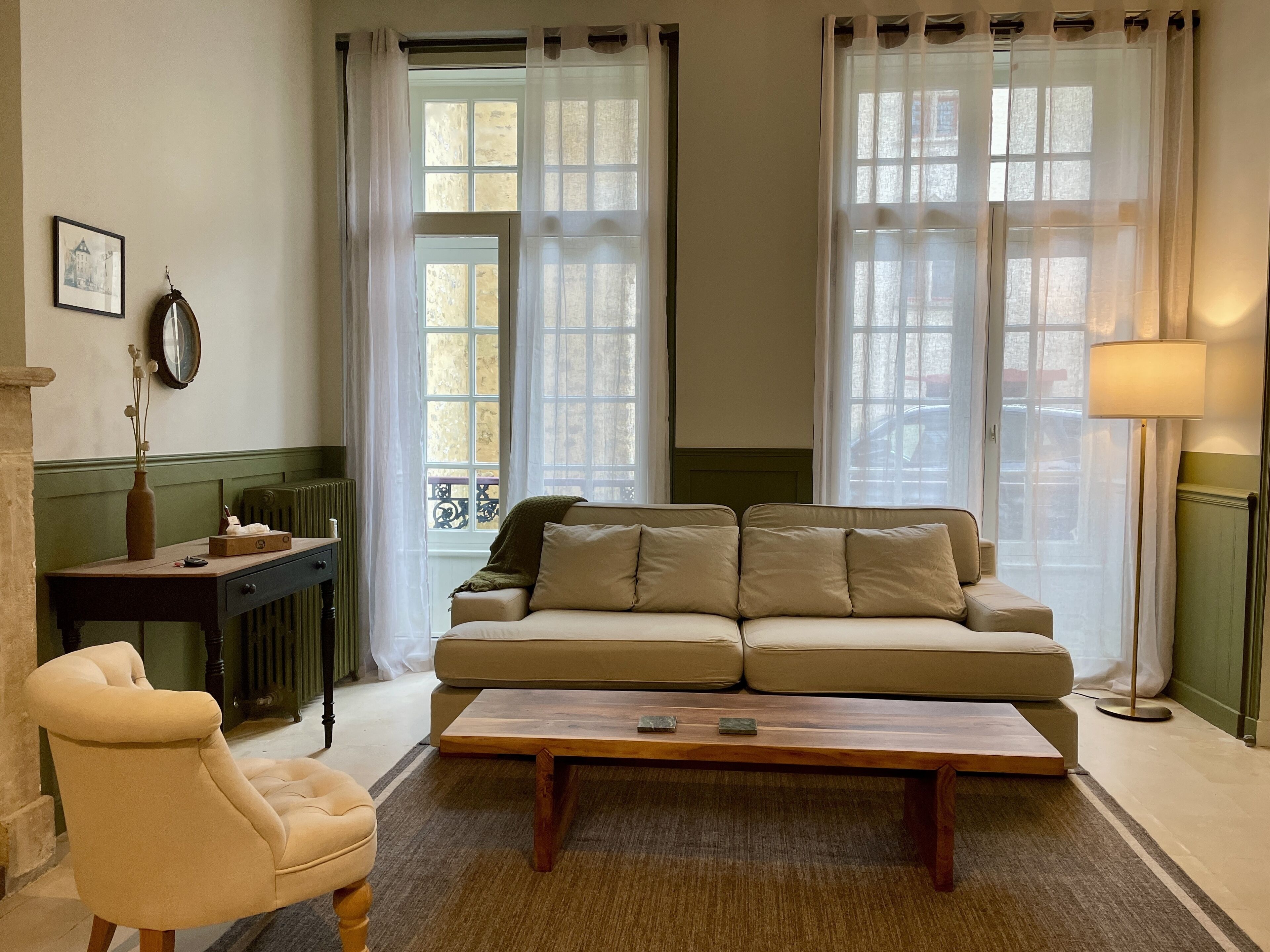 A living room with a sofa, armchair, and a coffee table. There are two windows with curtains, a radiator, and a lamp.