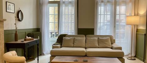 A living room with a sofa, armchair, and a coffee table. There are two windows with curtains, a radiator, and a lamp.