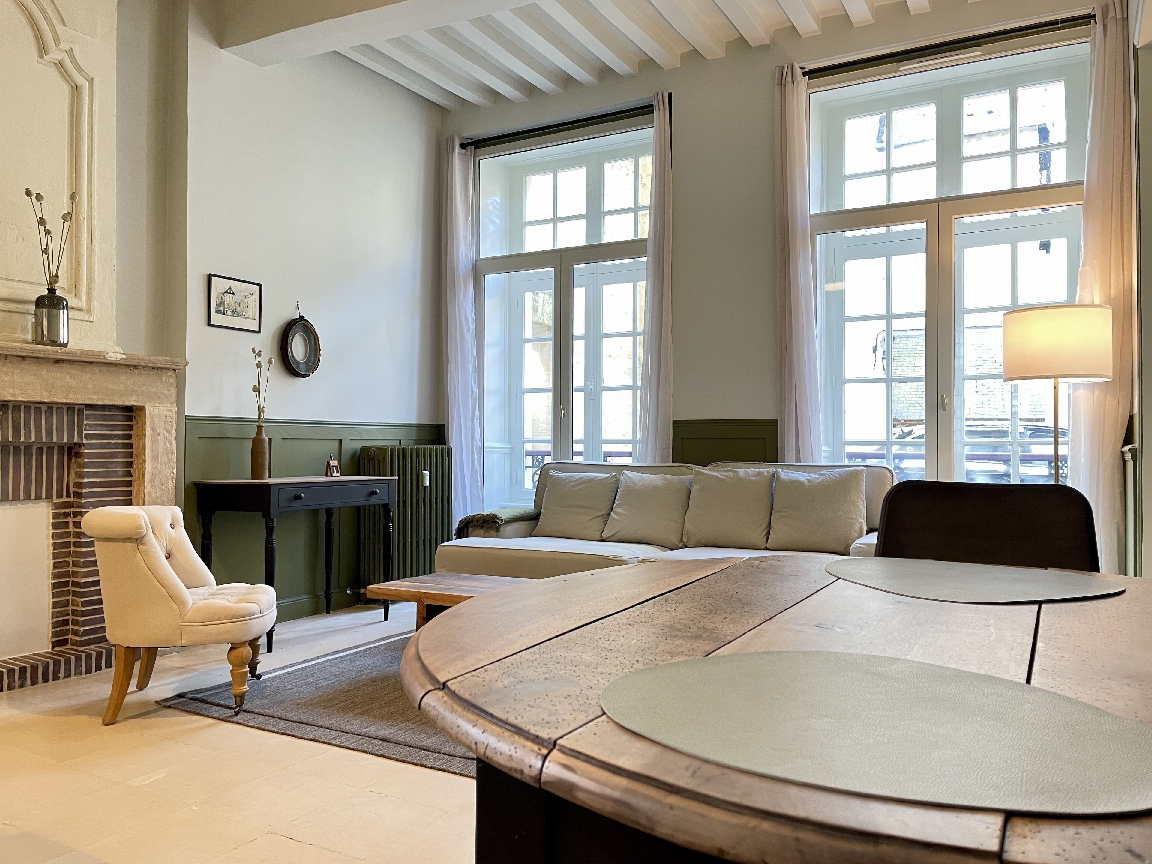 A room with a curved table, a sofa, a chair, a fireplace, and large windows.