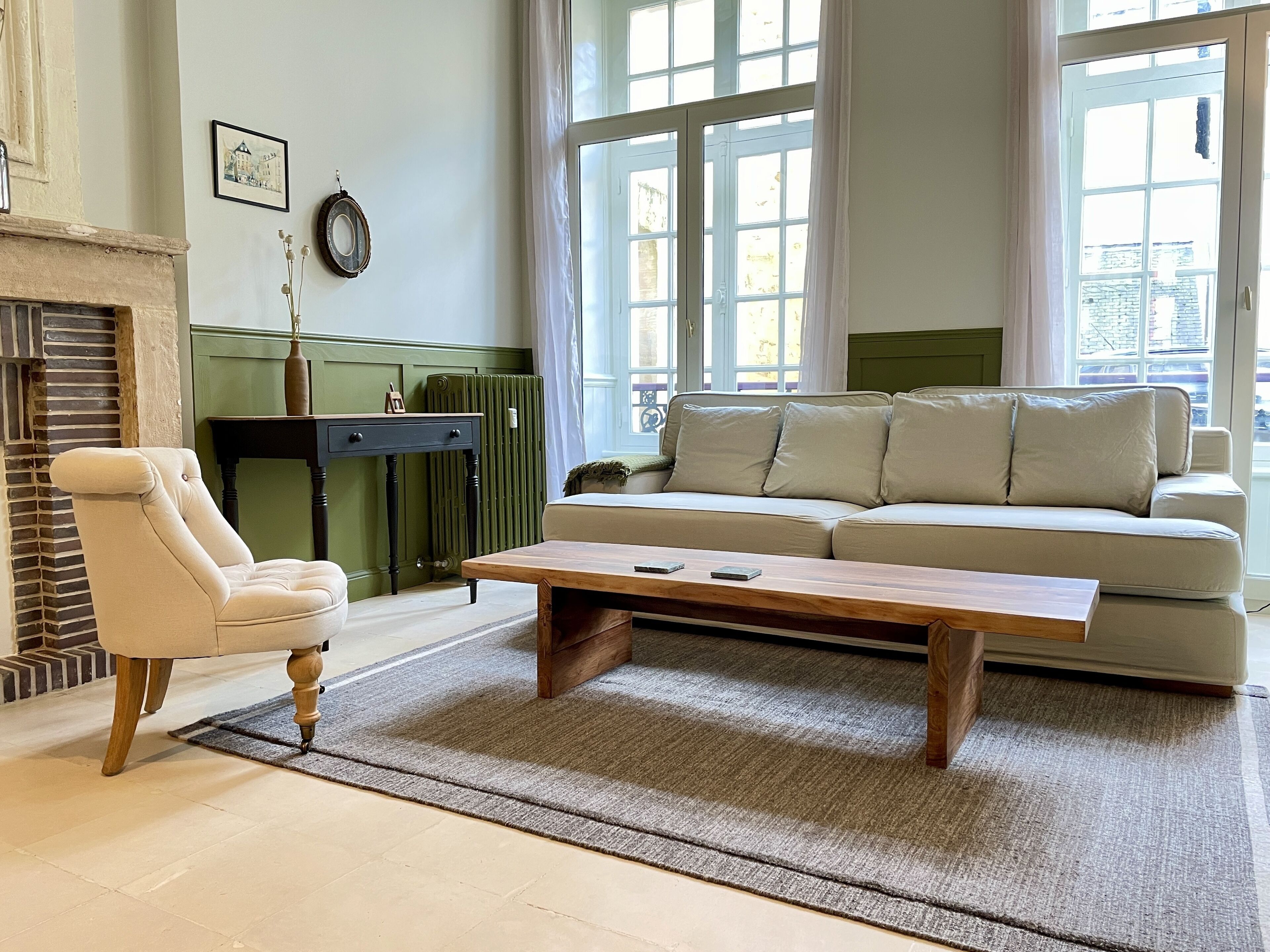 Uma sala de estar com sofá, poltrona e mesa de centro. Há um radiador, um quadro na parede e uma janela com cortinas.