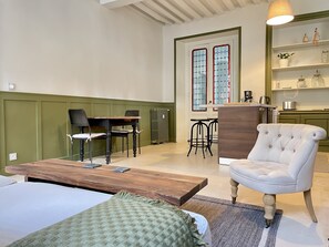 A room with a bed, a wooden coffee table, a white armchair, a dining table with chairs, and a kitchen area with a radiator and shelves.